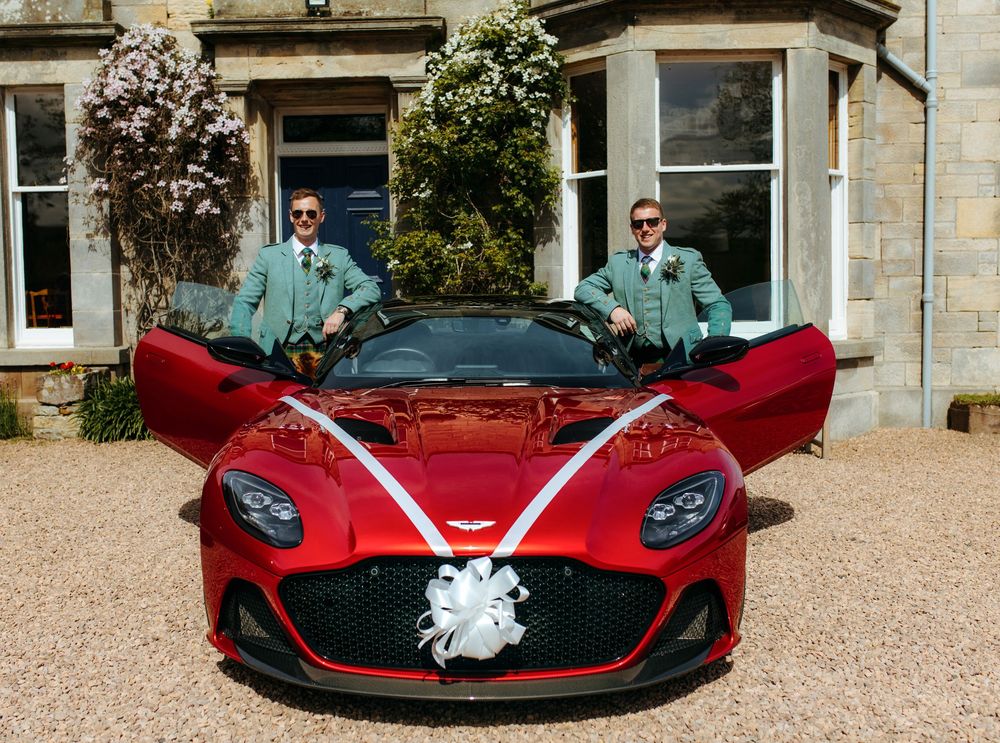 Two grooms in green suits stand beside a red Aston Martin sports car decorated with white wedding ribbons.