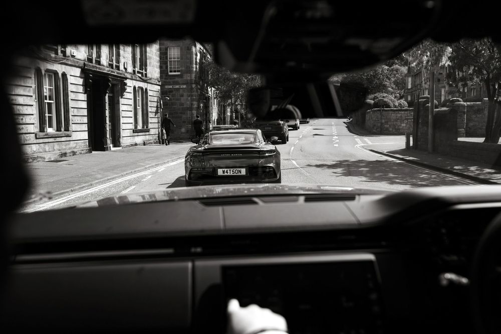 Black and white photo taken from inside a car, showing a classic sports car ahead on a quiet urban street.
