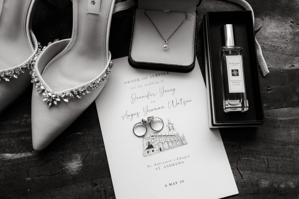 Black and white wedding flat lay with heels, invitation, rings, necklace, and Jo Malone perfume on dark wood.