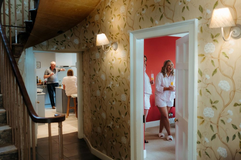 A woman in white poses in a bright red room while another person stands in a kitchen, seen through a floral wallpapered hallway.