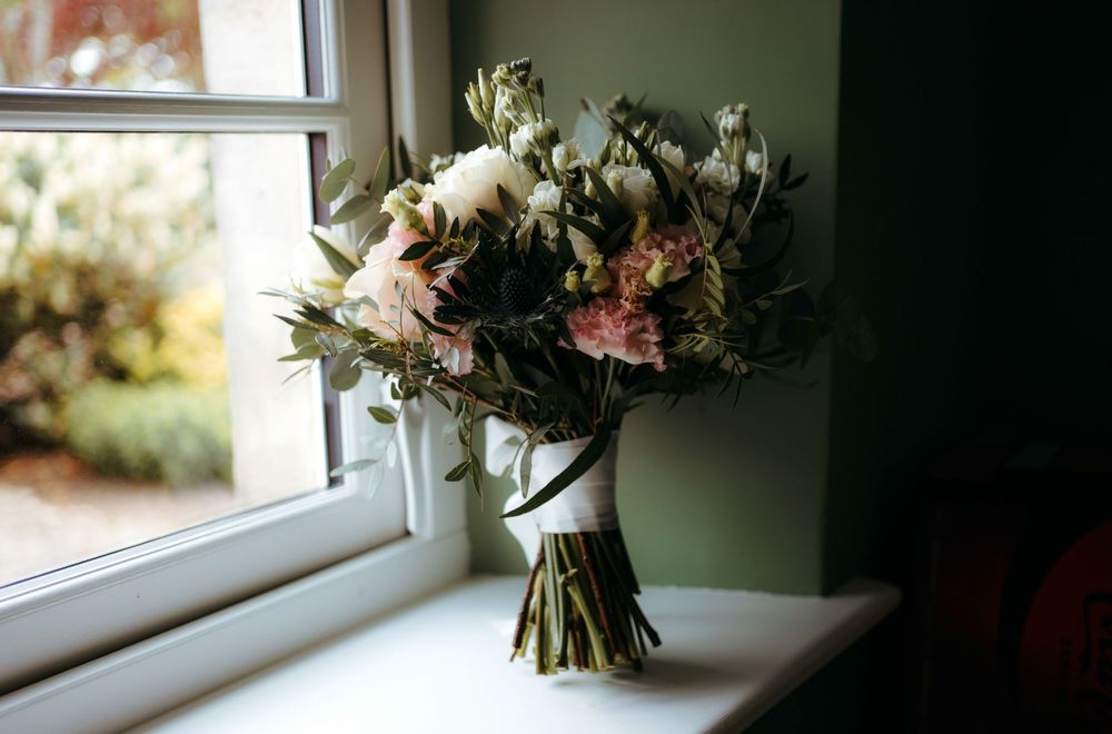 Elegant bridal bouquet with white, pink blooms and greenery resting on a windowsill in soft natural light.