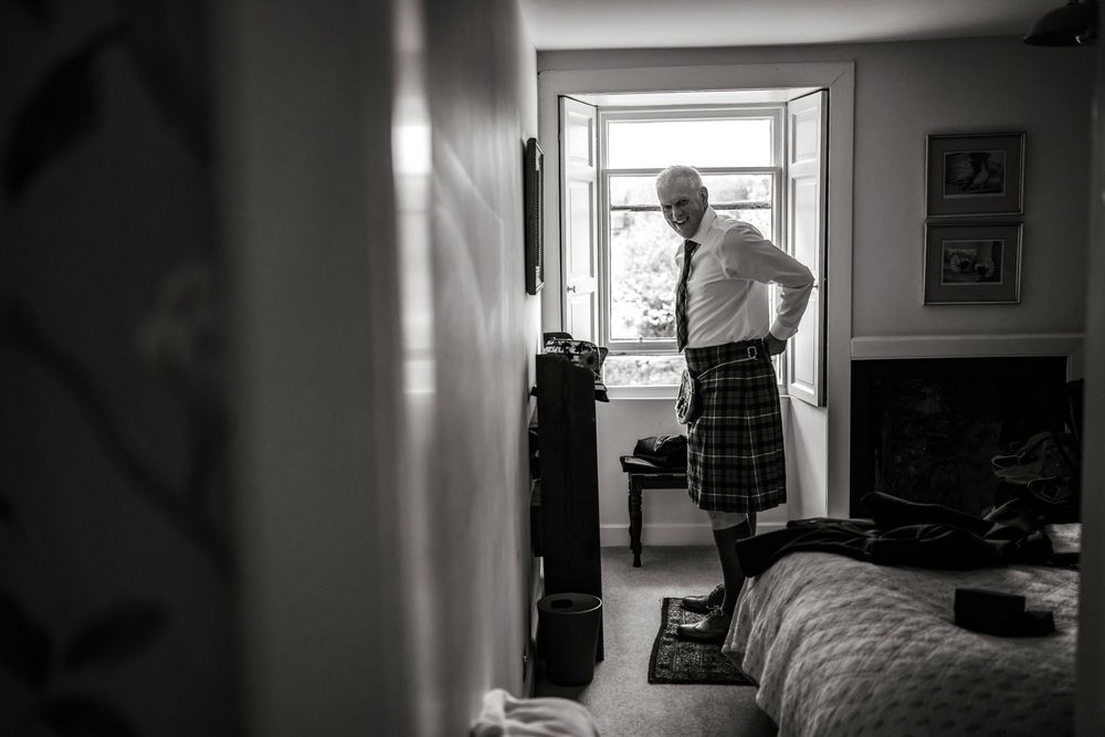 Black and white photo of a man in a kilt and tie standing by an open window in a bedroom.