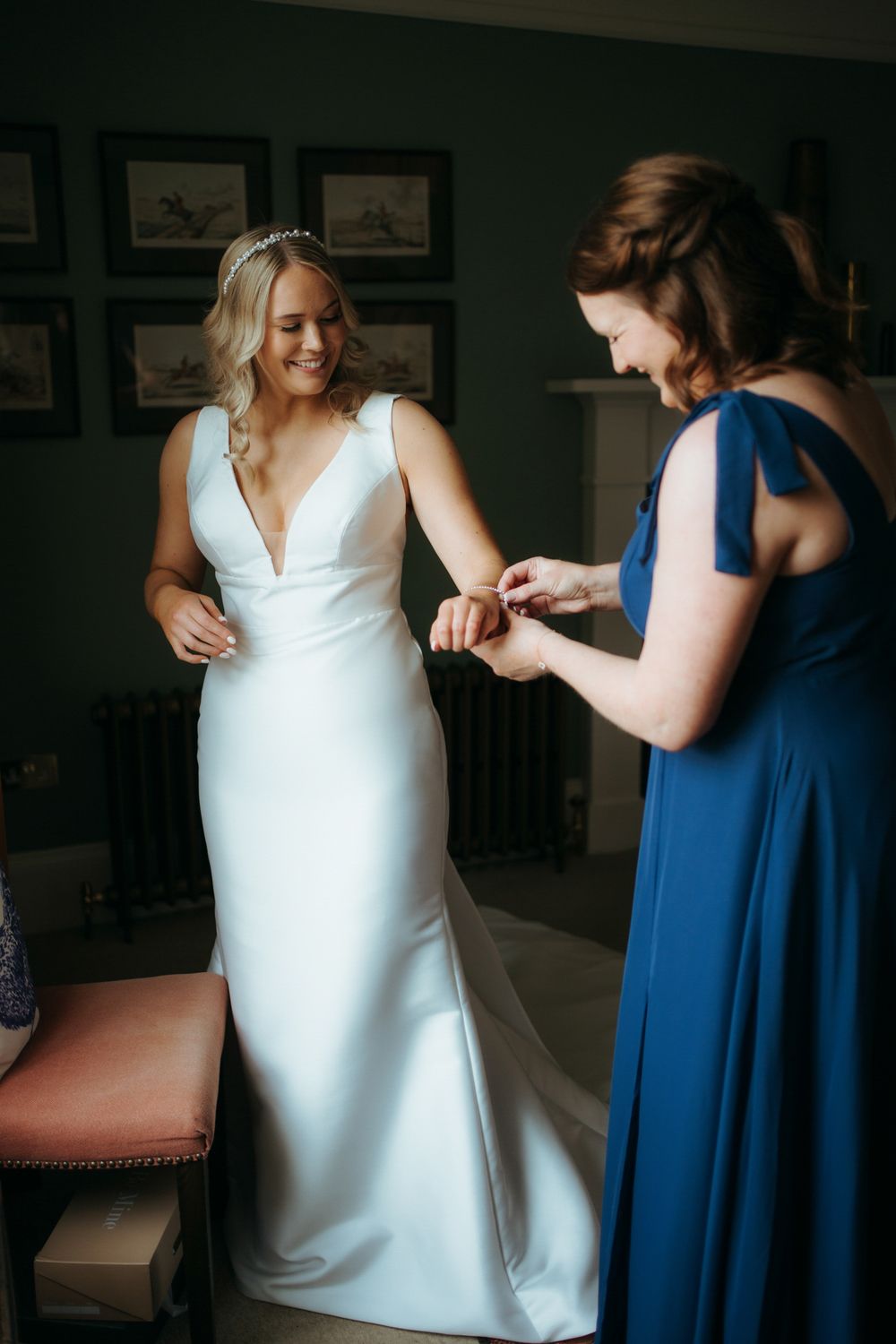 Bride in white gown smiles as bridesmaid in blue dress helps her with bracelet in elegant room.