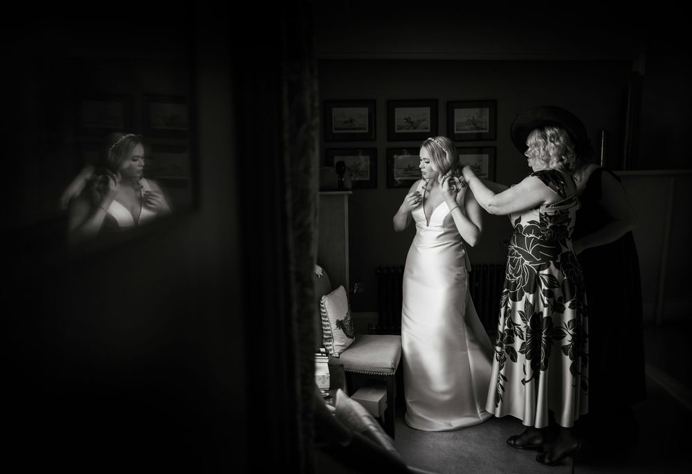 Black and white photo of a bride being helped into her wedding dress by two women in a softly lit room.