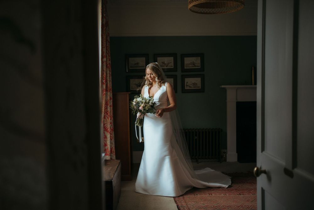 Bride in white gown holding bouquet stands in doorway of elegant room with soft natural light.