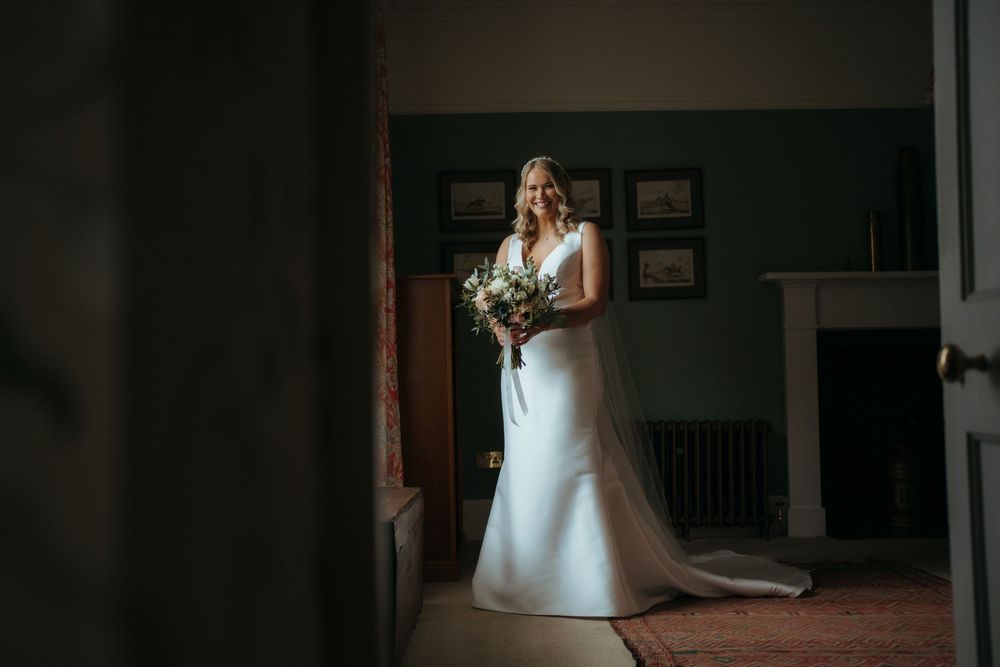 Bride in white gown holding bouquet stands in elegant room with fireplace and green walls on her wedding day.