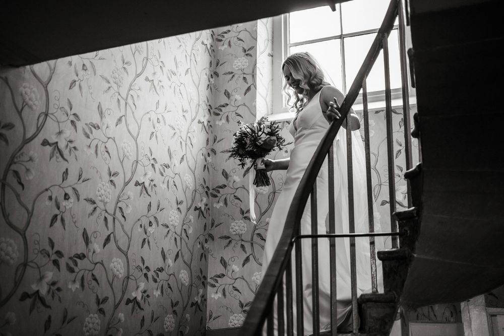 Bride in white dress holding bouquet descends elegant spiral staircase with floral wallpaper in black and white photo.