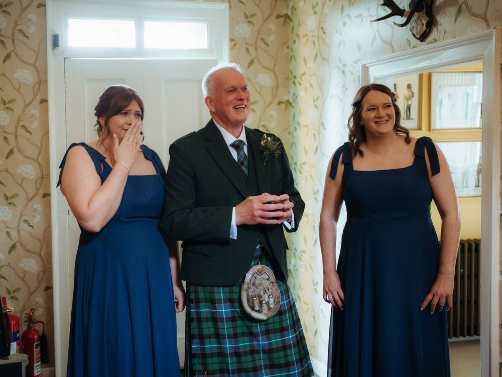 Man in Scottish kilt with two women in blue dresses laughing together at a wedding celebration.