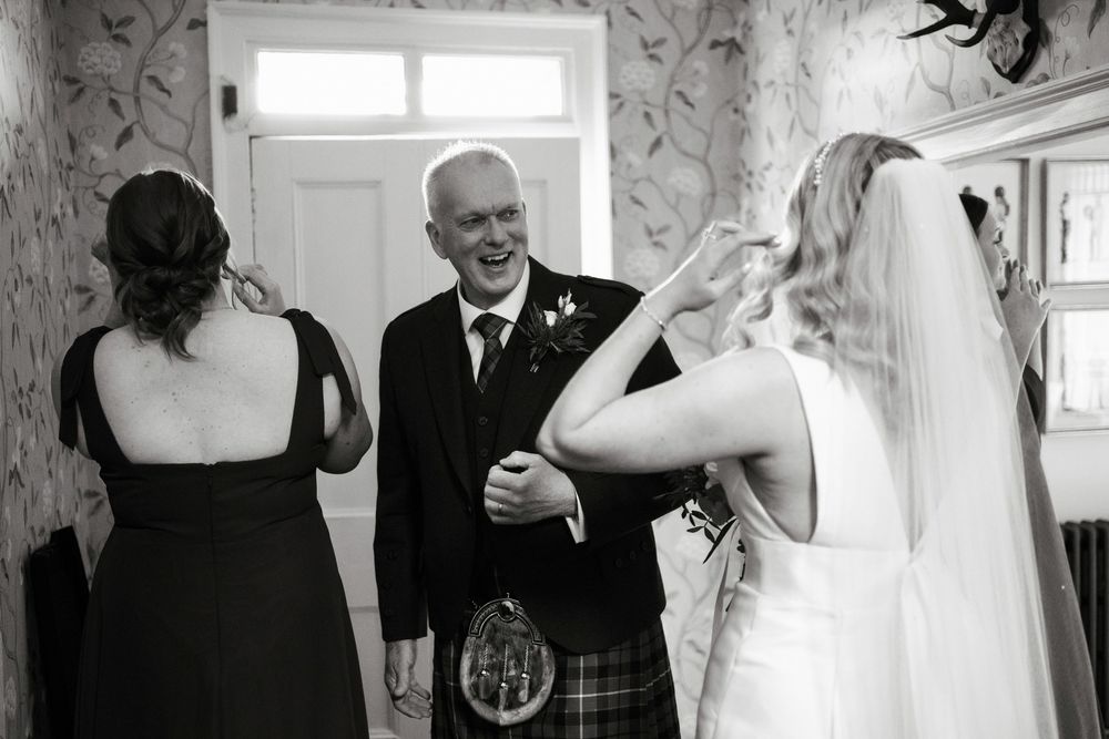 Black and white photo of a bride in veil meeting smiling man in Scottish kilt tartan at a wedding celebration indoors.
