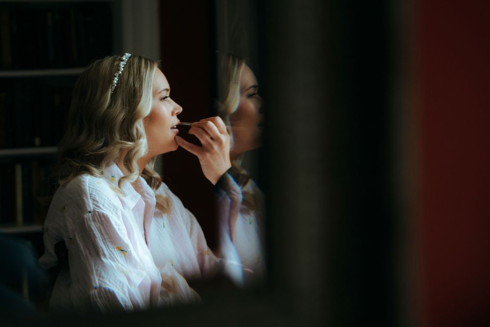 Bride applying makeup in mirror reflection wearing white robe and hair accessory during wedding preparation.