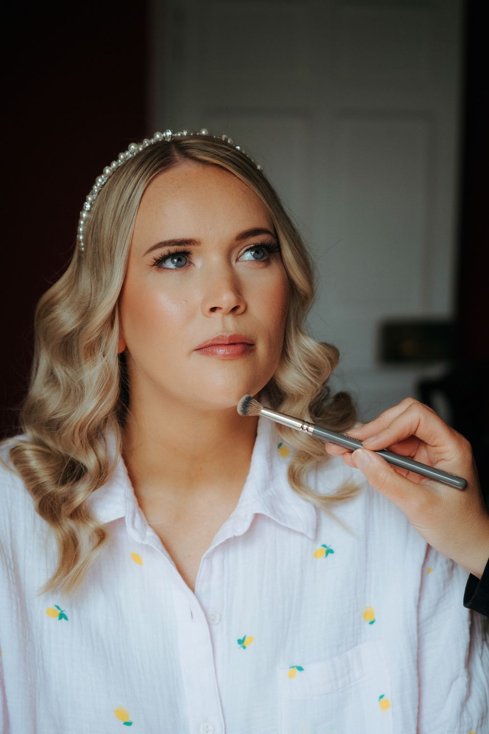 Bride getting makeup applied with a brush, wearing a pearl headband and white floral shirt, with soft curled blonde hair.