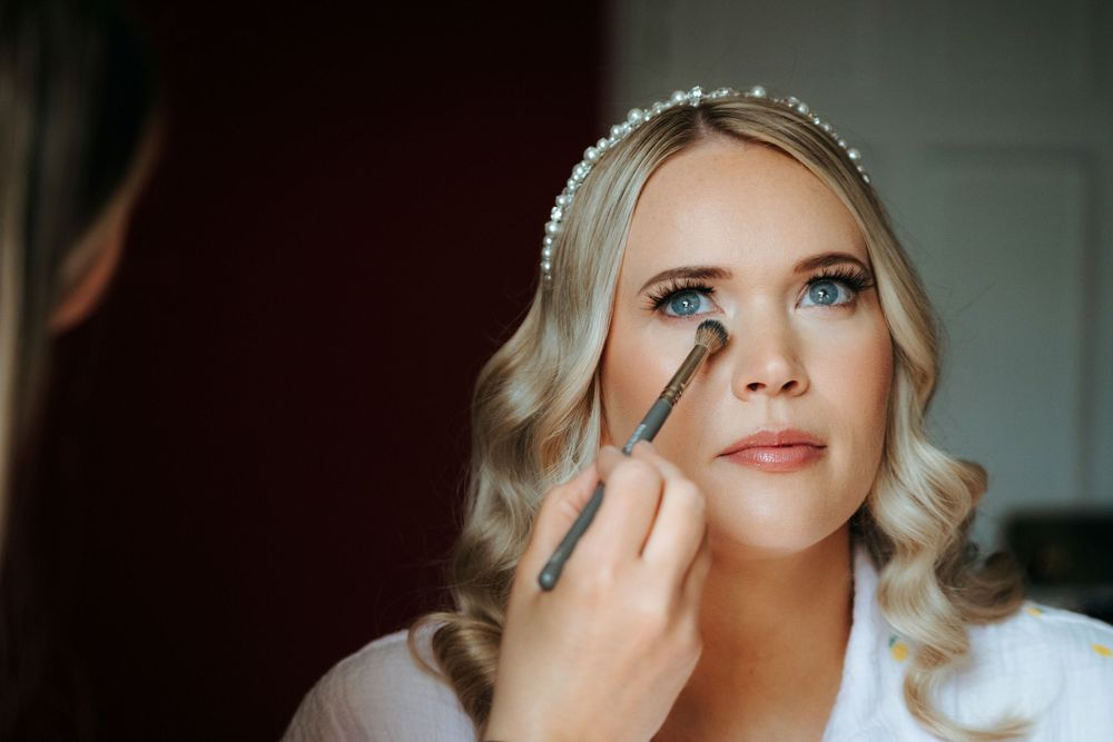 Bride getting eye makeup applied with a brush, wearing a pearl headband, with soft curled blonde hair.