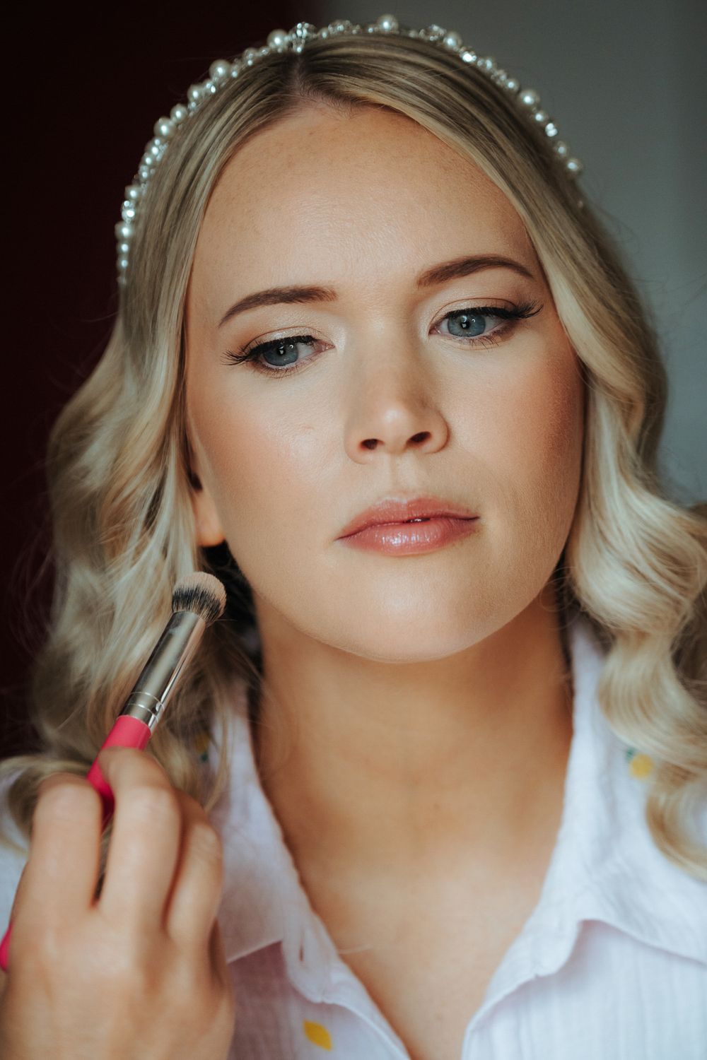 Blonde bride wearing pearl headband gets makeup applied with brush during bridal beauty preparation.