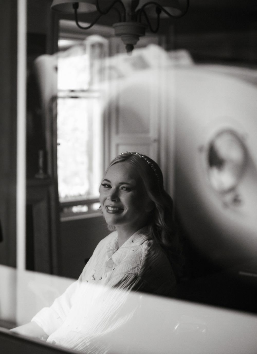 Smiling bride in a white robe with pearl headband reflected in a mirror in black and white photo.
