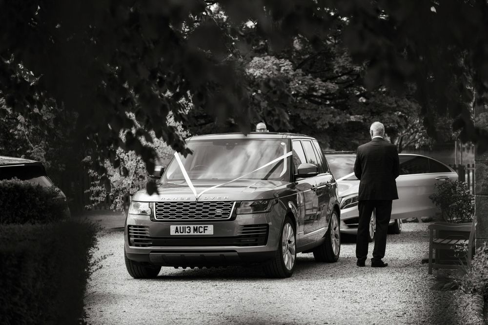 Black and white photo of a Range Rover decorated with white ribbons on a gravel driveway at a wedding.