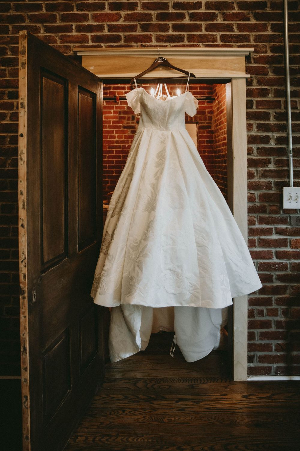 Elegant white ball gown wedding dress hanging on a wooden door frame against a rustic brick wall backdrop.