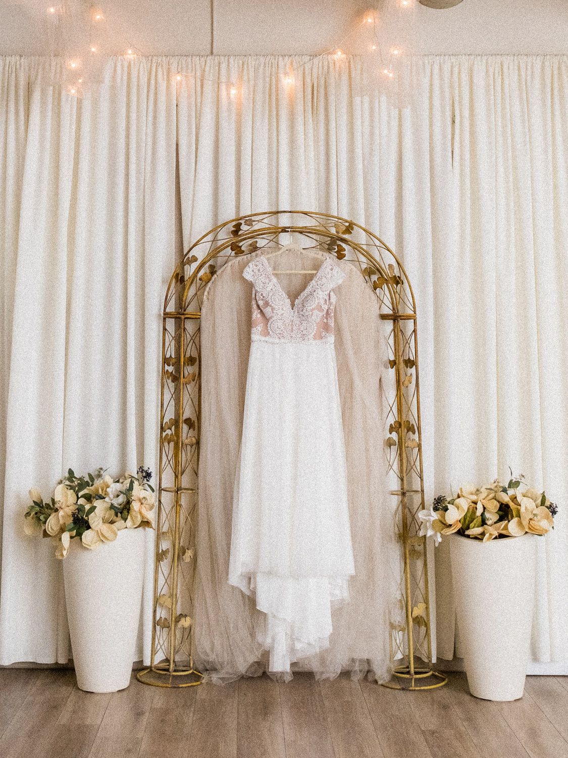 White wedding dress hanging on gold arch with floral arrangements, crystal chandelier, and fairy lights in elegant bridal suite.