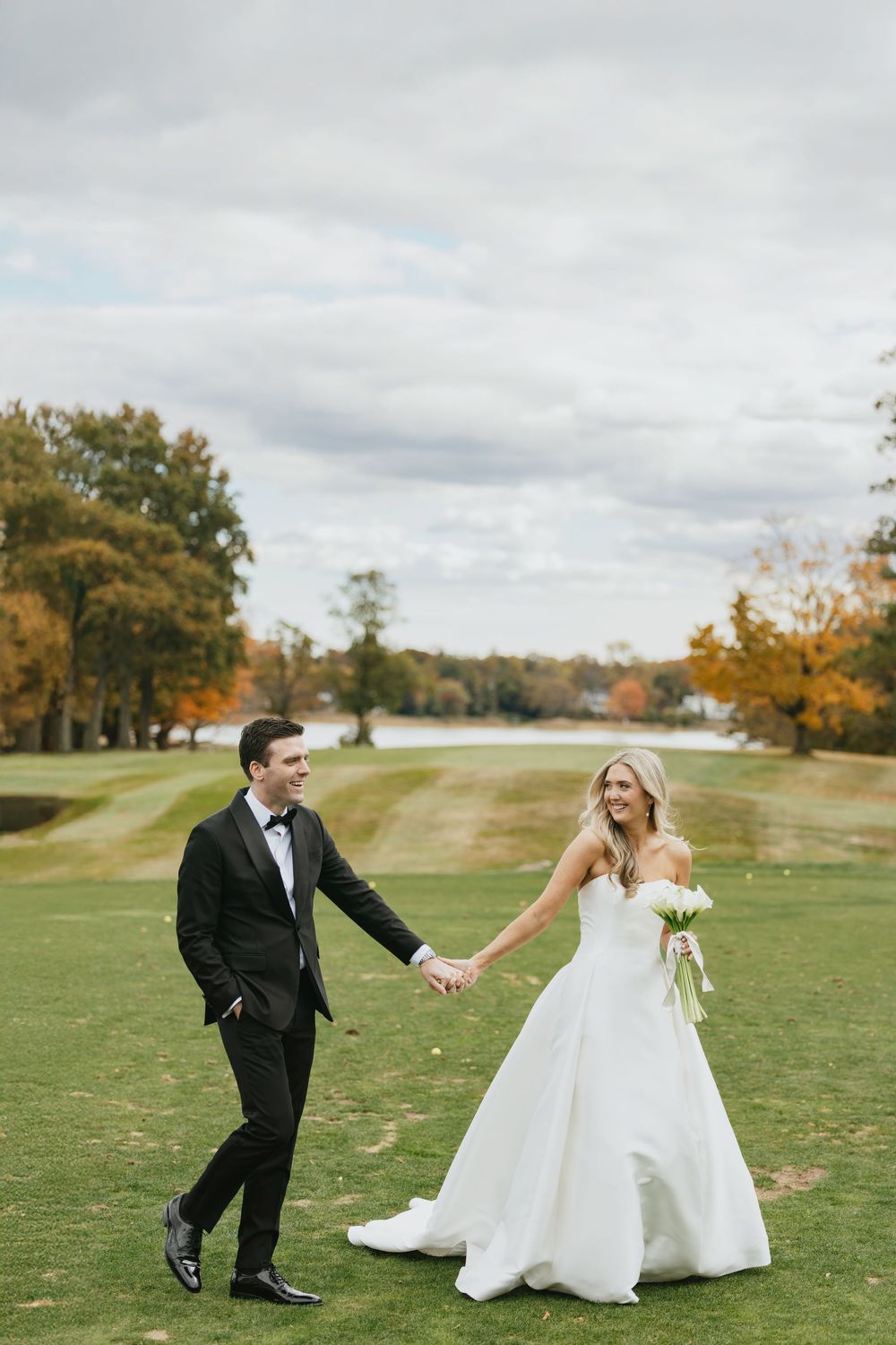 A bride and groom hold hands walking on a green golf course surrounded by autumn trees on their wedding day.