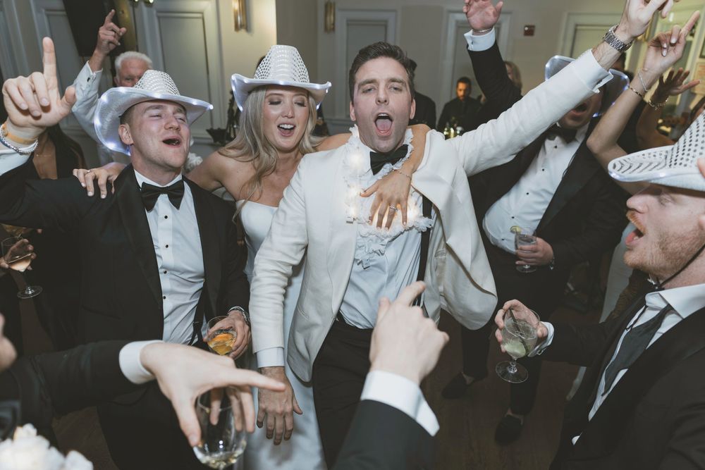 Joyful wedding party celebrating in cowboy hats and tuxedos, raising glasses and dancing at a lively indoor reception.