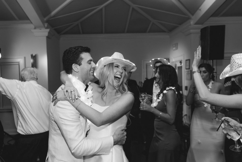 Black and white photo of a joyful bride in cowboy hat dancing with groom at lively wedding reception.
