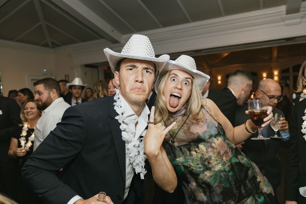 Two people wearing white cowboy hats pose playfully at a festive indoor party event.