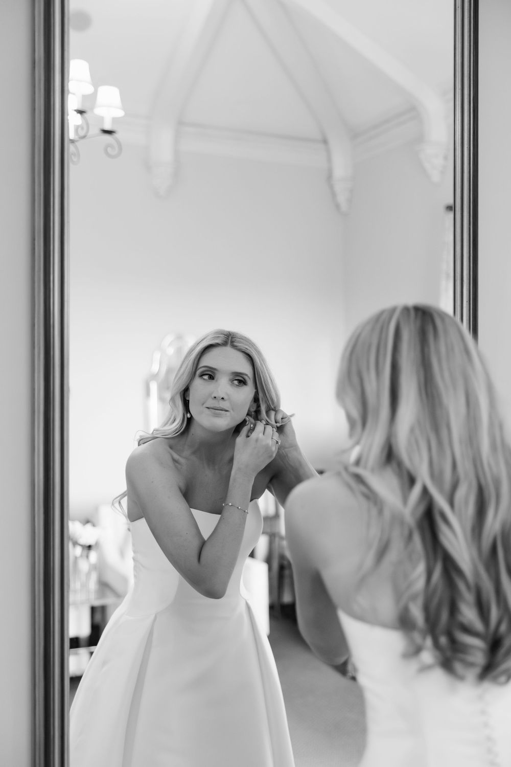 Bride in white gown adjusting earring while looking in mirror, elegant black and white wedding photo.