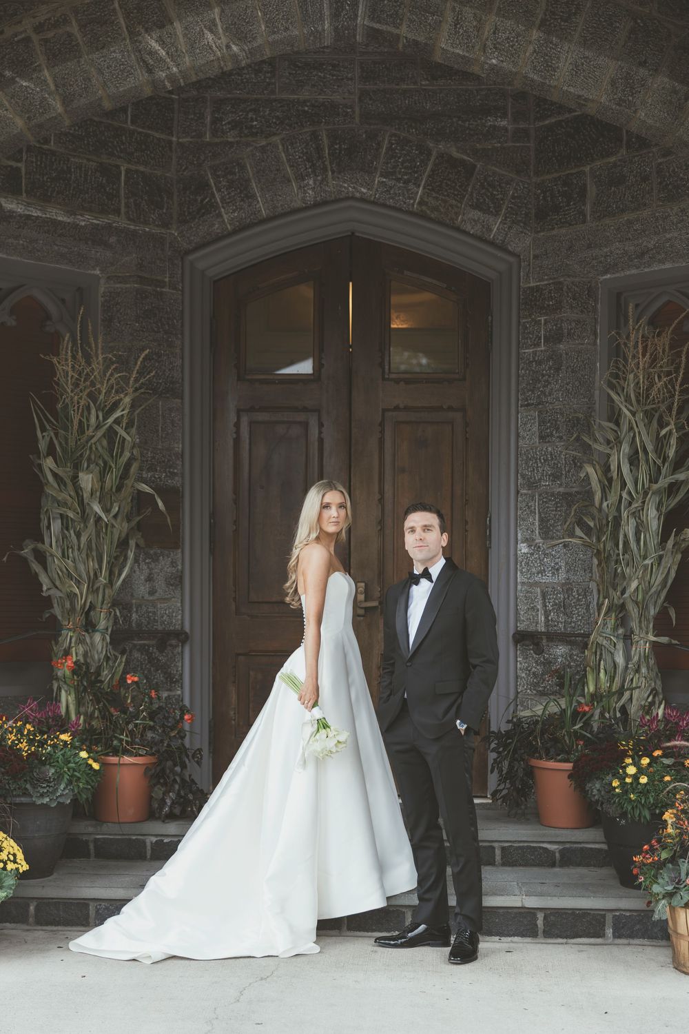 Bride in white gown and groom in black tuxedo pose before ornate stone church entrance with wooden doors.