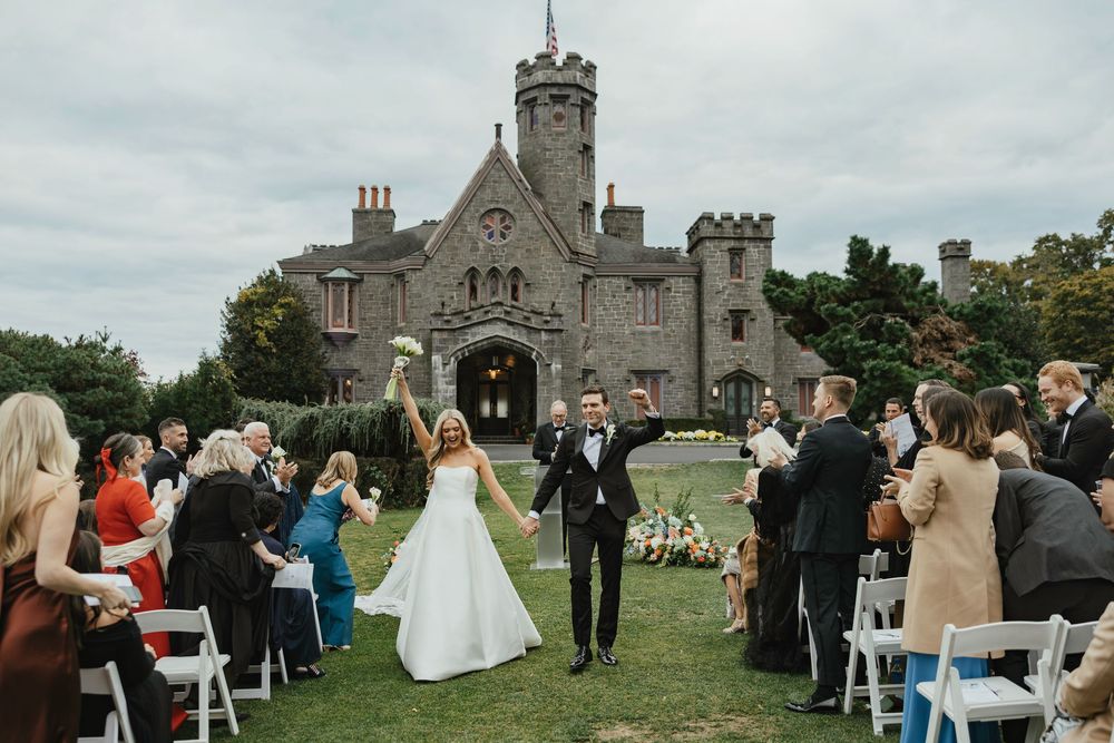 Newlyweds celebrate recessional outside a historic stone castle venue with guests cheering on a cloudy day.