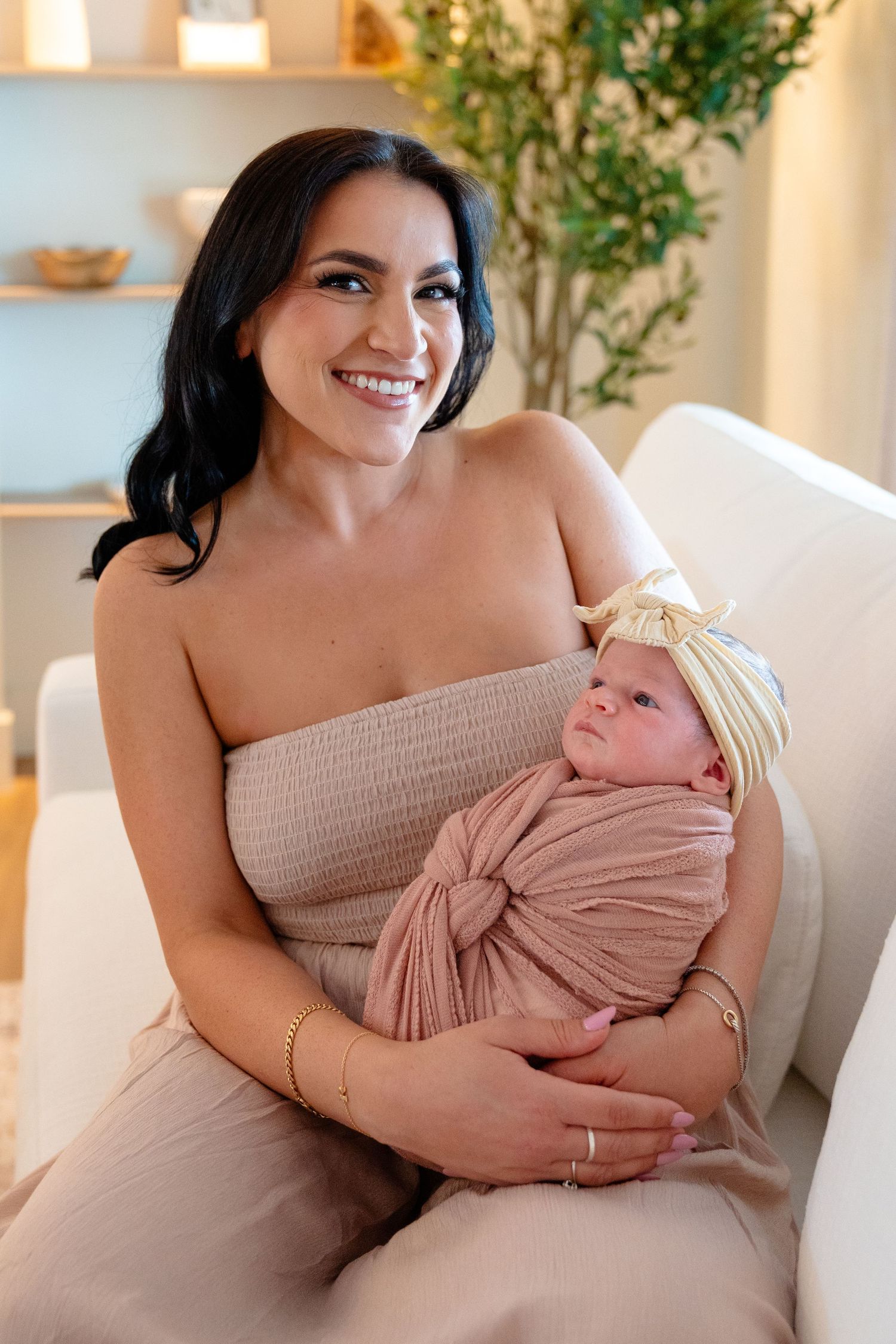 Smiling brunette mother holding swaddled newborn baby in pink wrap and yellow bow headband on white chair.