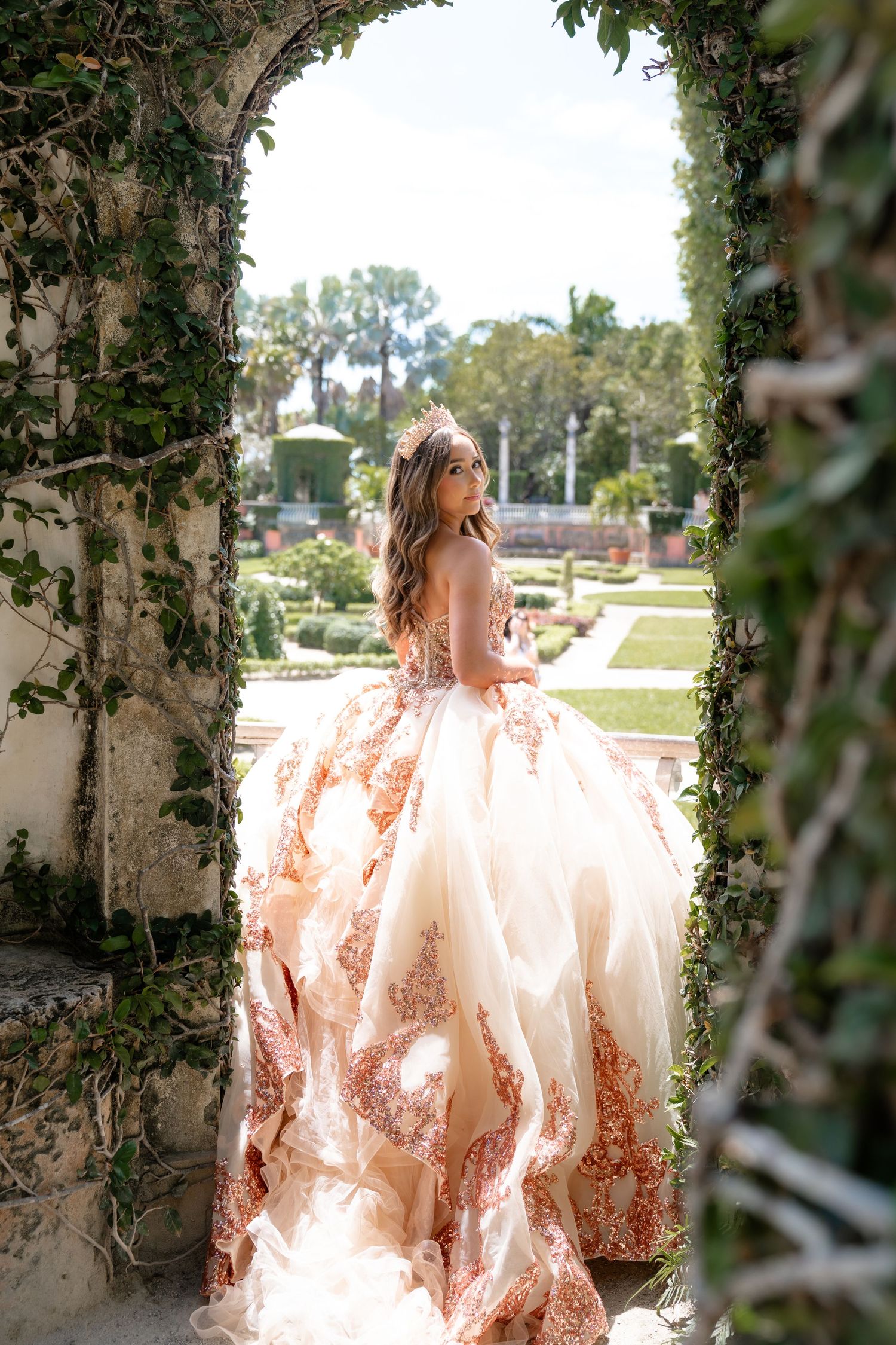 Young woman in an ornate rose gold ballgown stands in an ivy-covered stone archway overlooking a lush formal garden.