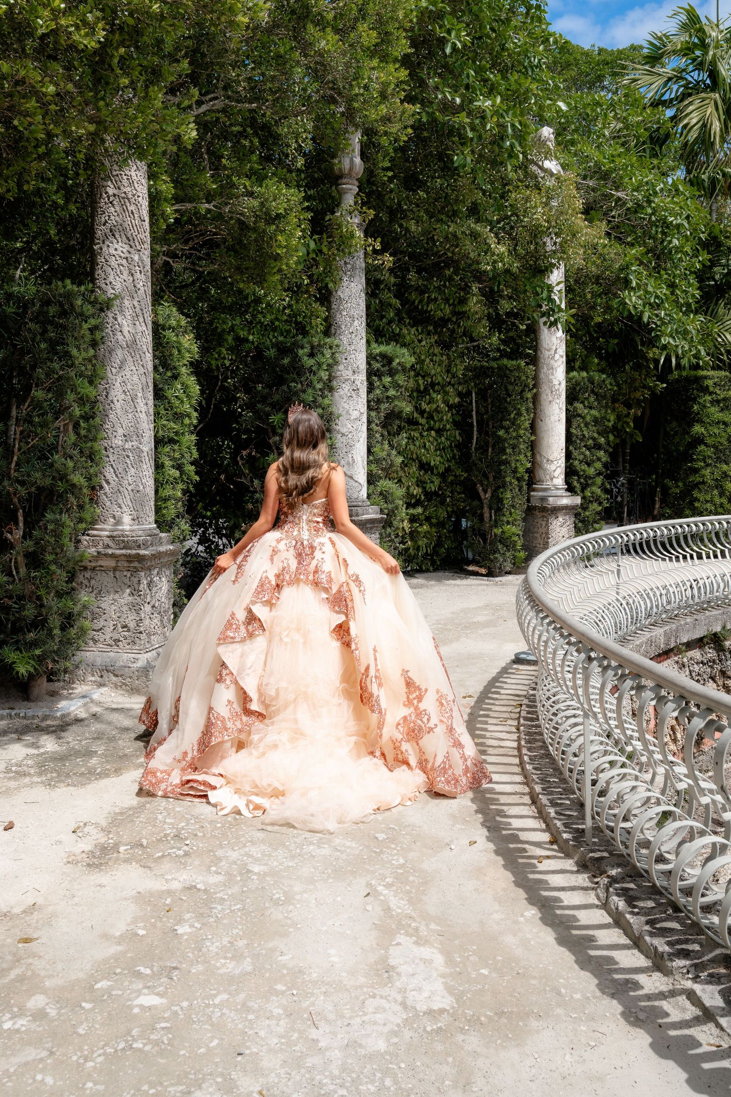 Woman in ornate ballgown with floral details stands on stone path surrounded by tall columns and lush greenery.