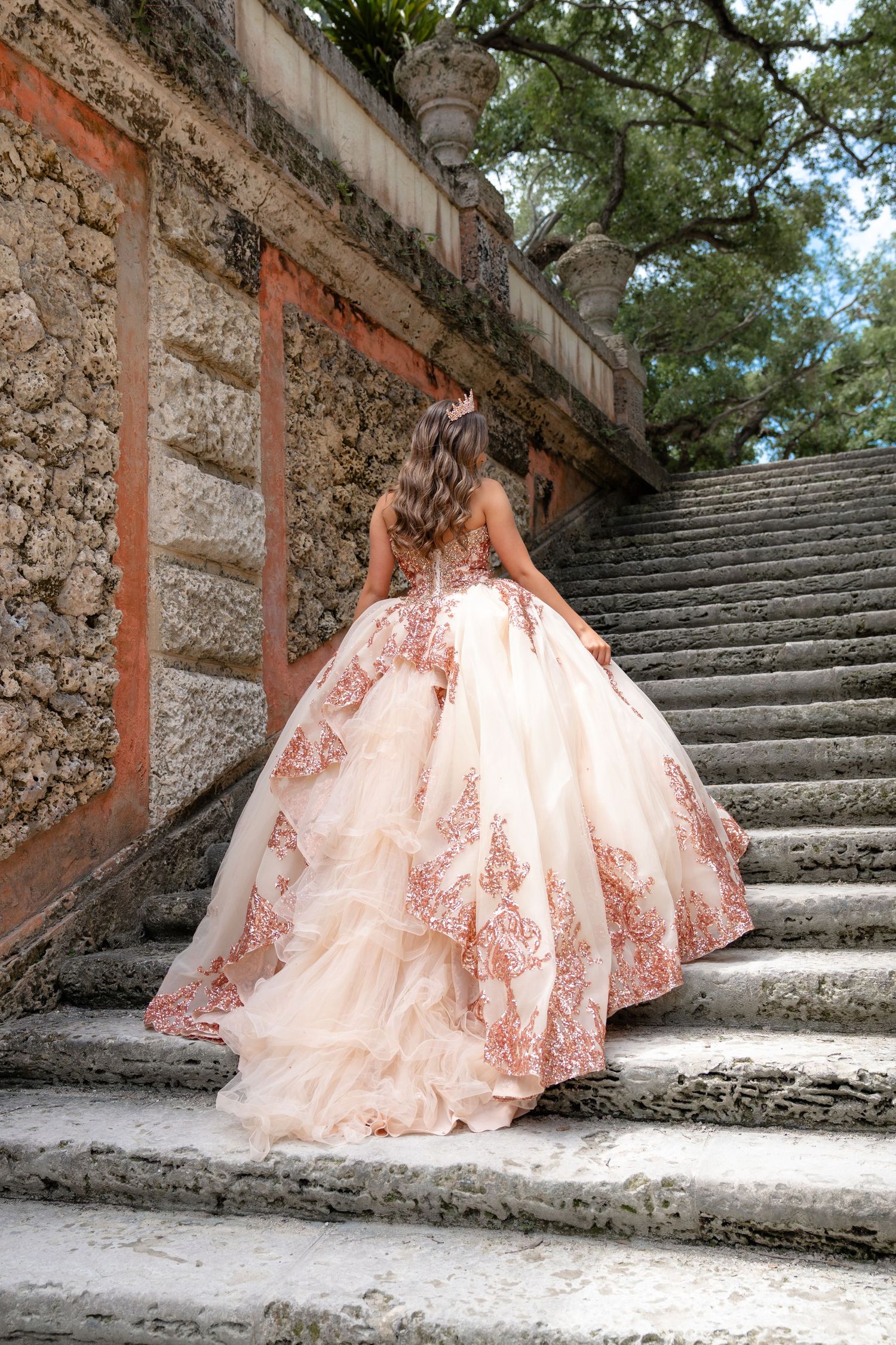 Young woman in a stunning blush pink ballgown with rose gold lace ascends stone stairs at an elegant outdoor venue.