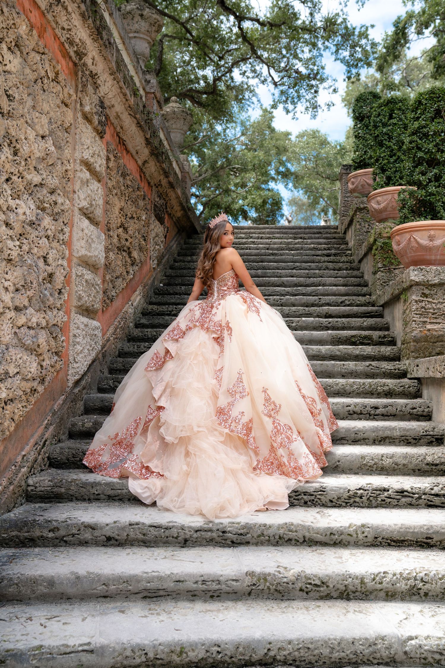 Young woman in a blush pink ball gown with rose gold lace ascending stone steps in a garden.