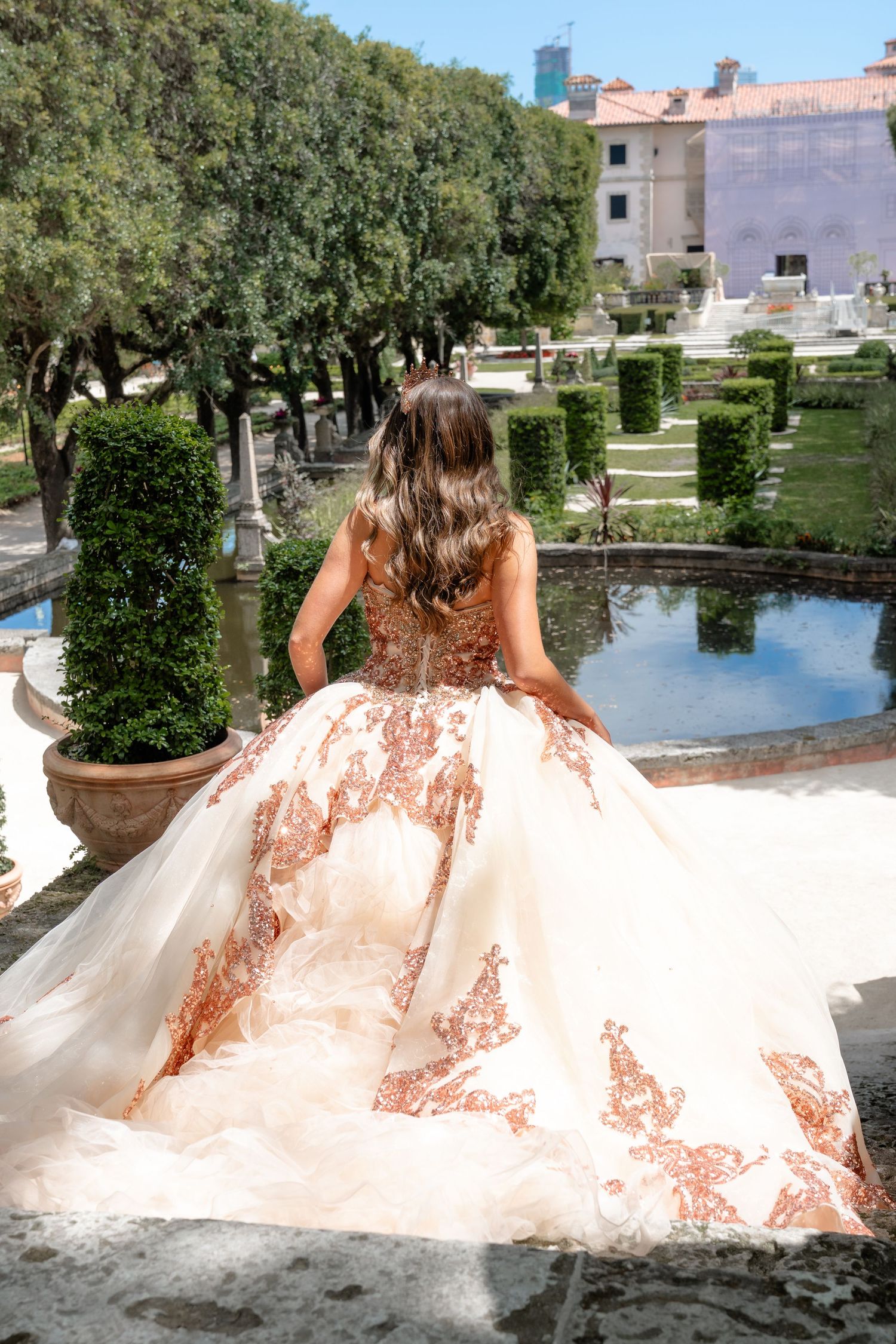 Young woman in elegant white and rose gold ball gown poses in formal garden near fountain.