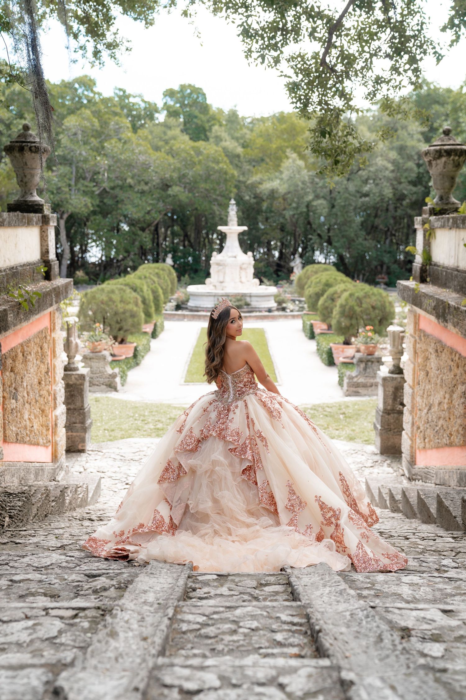 Elegant quinceañera in a blush ruffled ball gown posing on stone steps with a fountain garden backdrop.