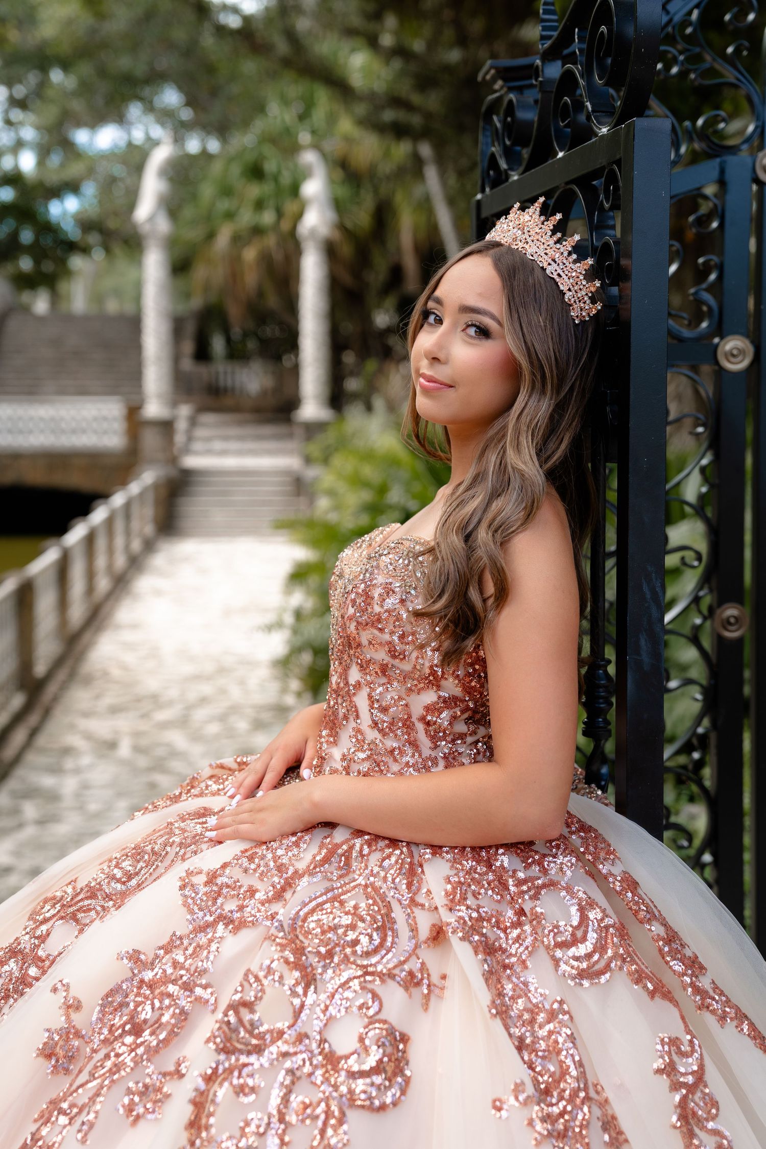 Young woman in rose gold sequin ball gown and tiara poses elegantly by ornate iron gate for quinceañera.