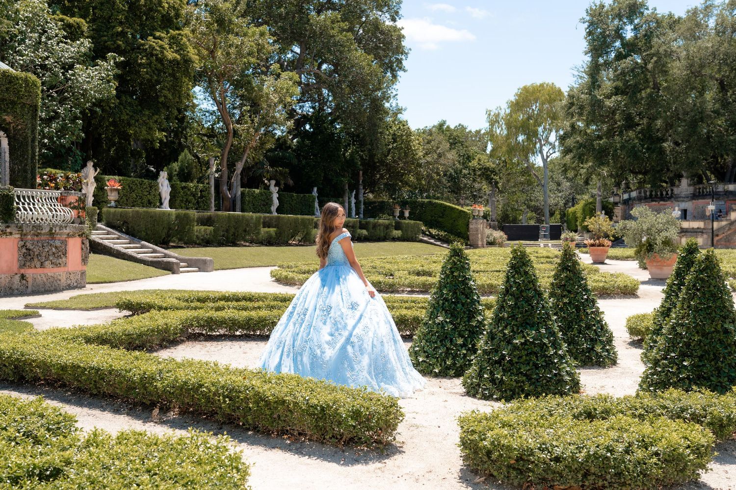 Young woman in a blue ball gown poses in a formal garden with trimmed hedges and cone-shaped topiaries.