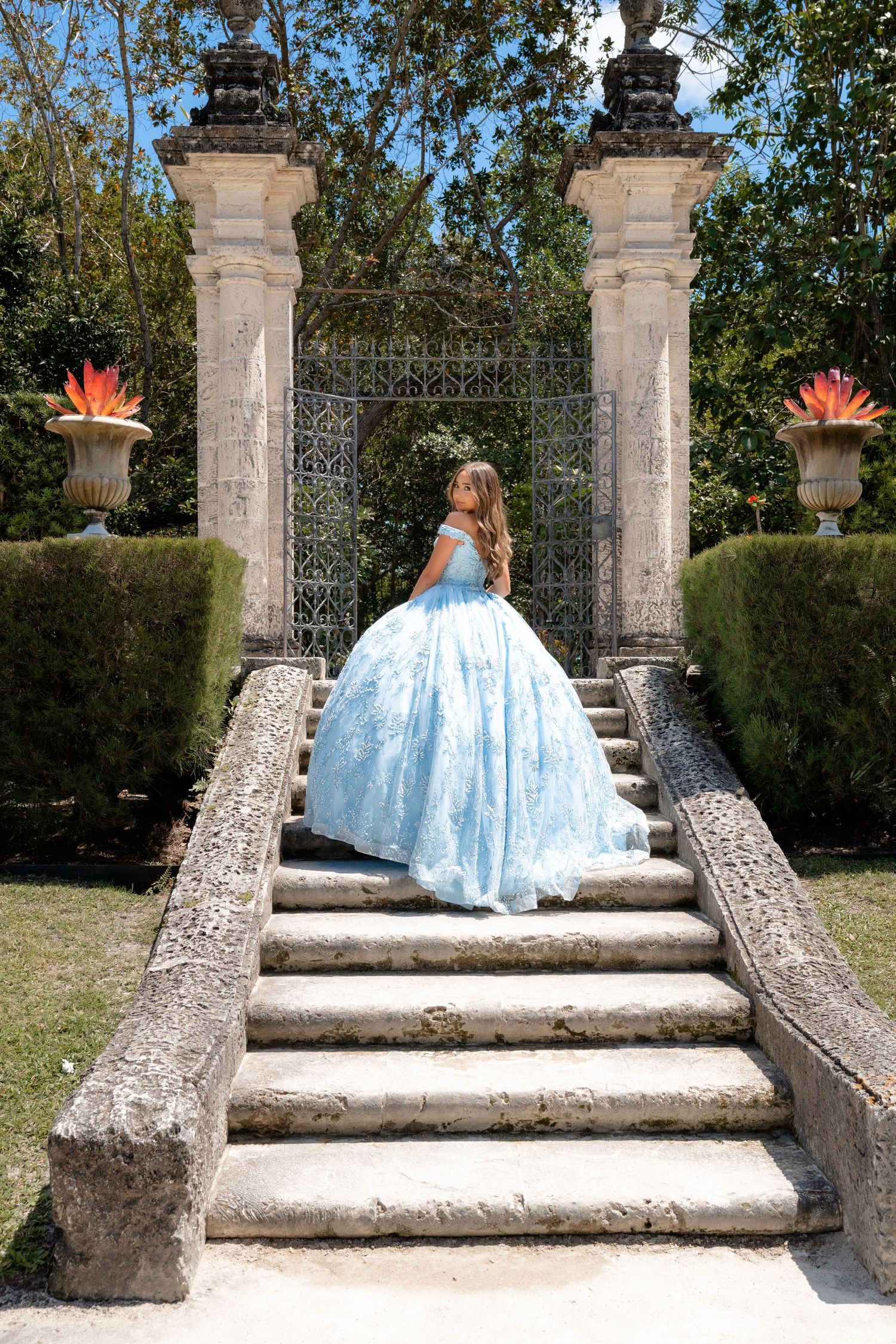 Young woman in a light blue ball gown ascending stone steps at an ornate garden gate.