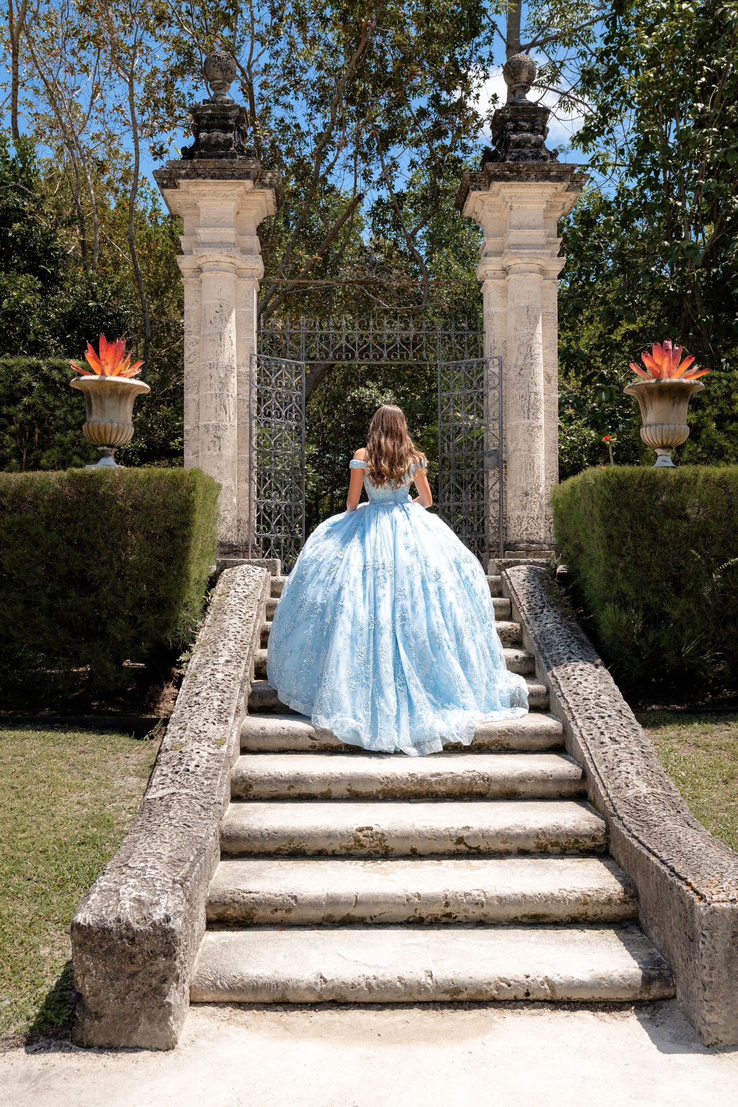 Girl in blue ball gown ascending stone steps toward ornate garden gate.