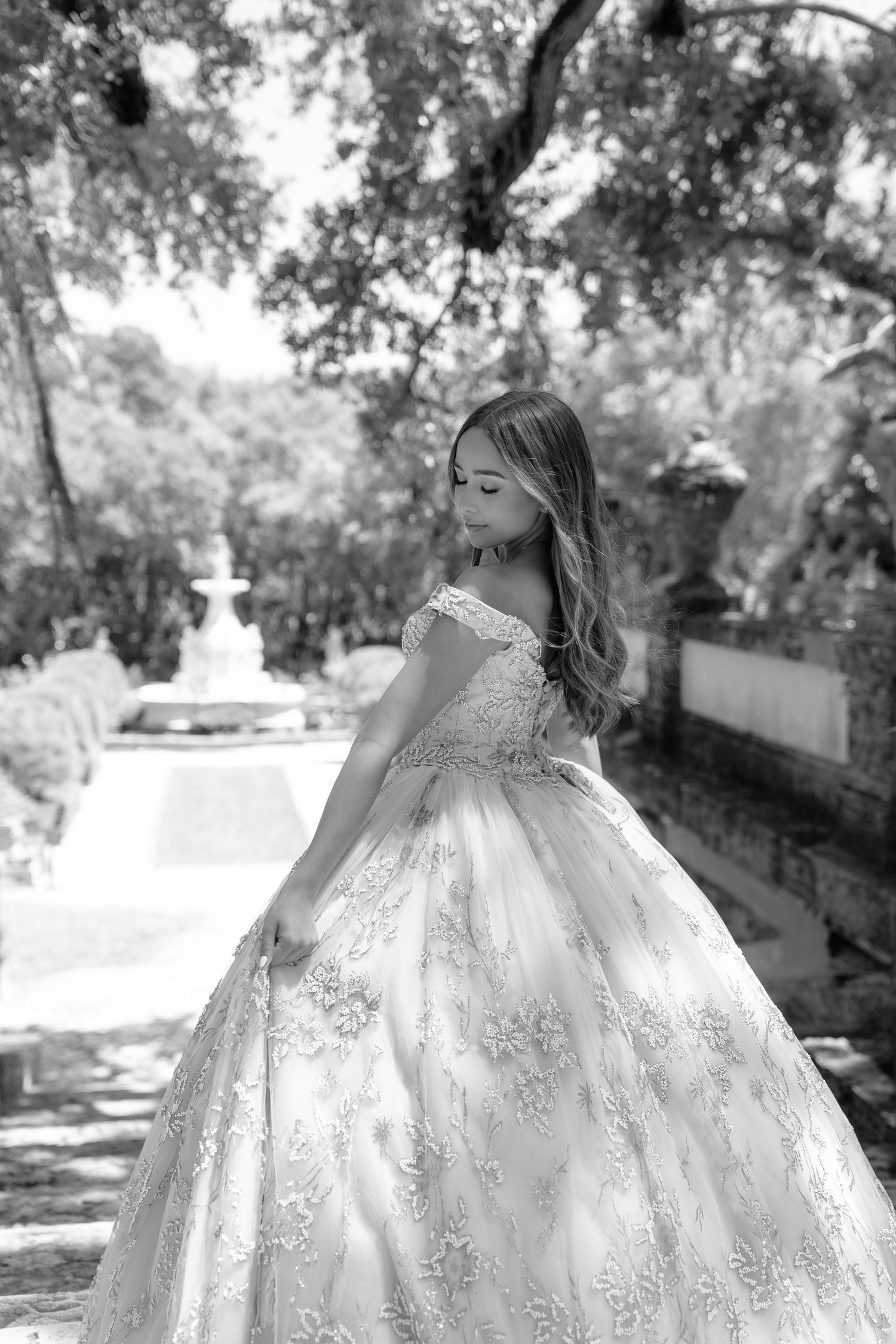 Young girl in elegant lace ball gown poses gracefully in a garden setting with fountain backdrop, black and white photo.