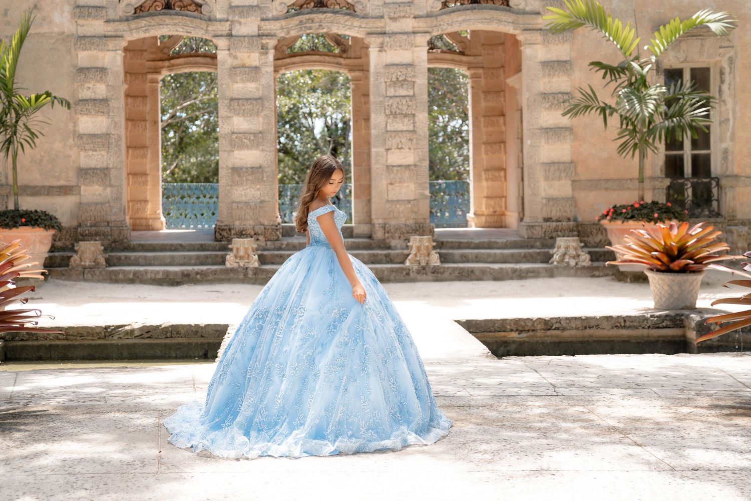 Young girl in a light blue ball gown quinceañera dress poses in an elegant outdoor courtyard setting.