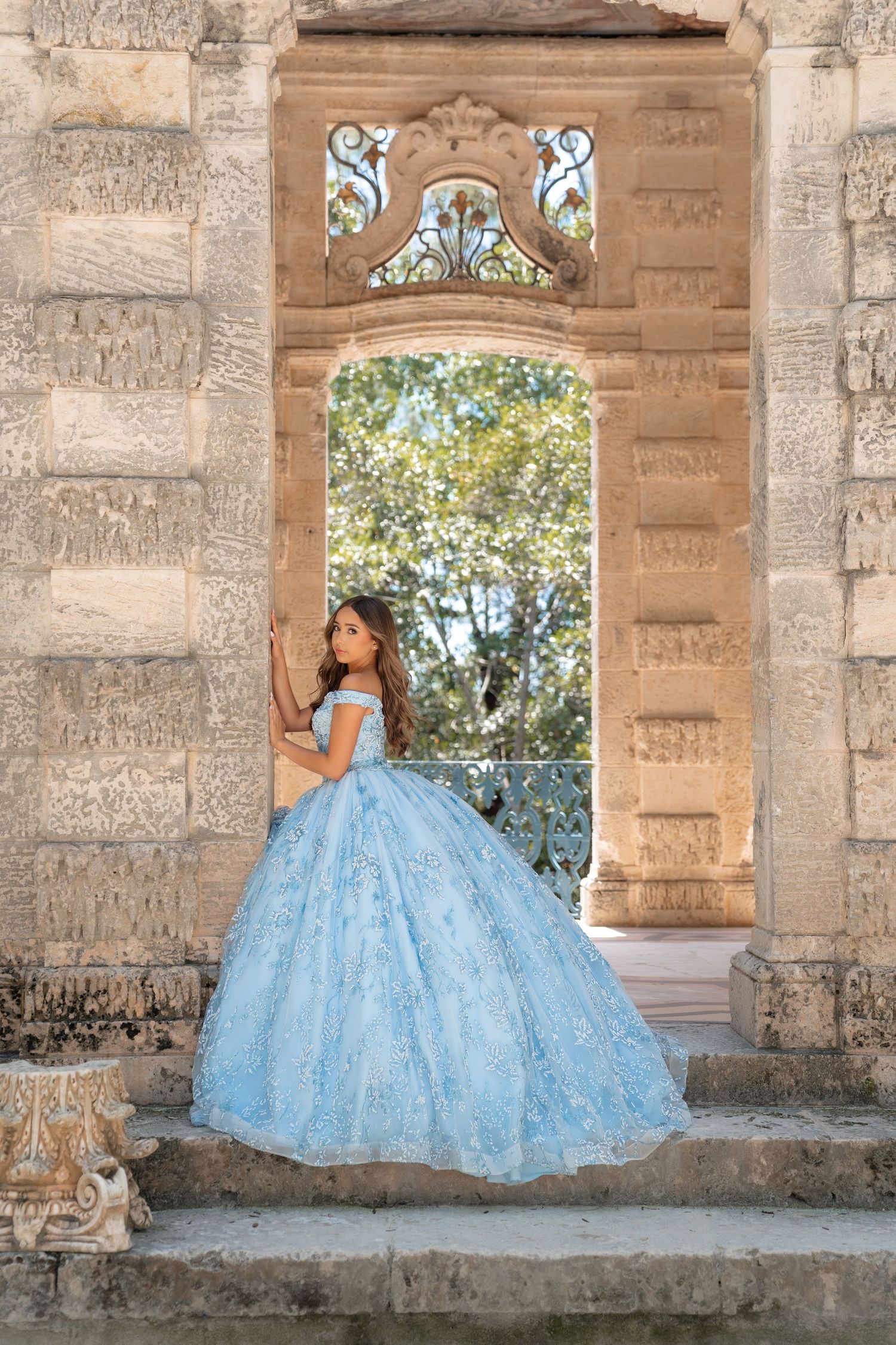 Young woman in a light blue ball gown quinceañera dress posing on stone steps of an ornate archway.