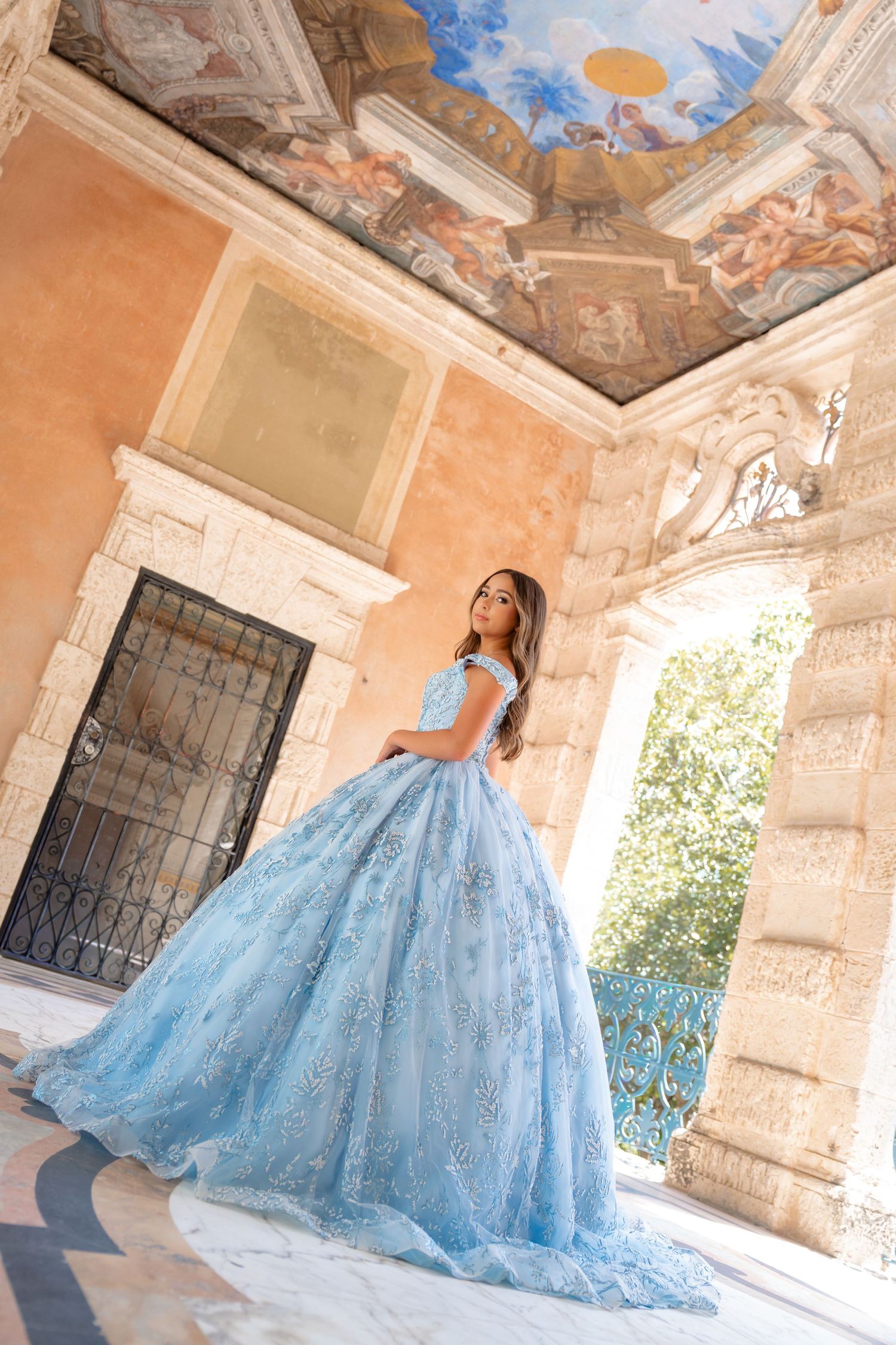 Young woman in a blue ball gown poses in an ornate building with a painted ceiling and marble columns.