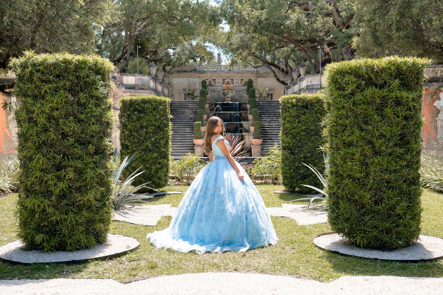 Young woman in a blue ball gown poses in a formal garden with tall hedges and a fountain backdrop.