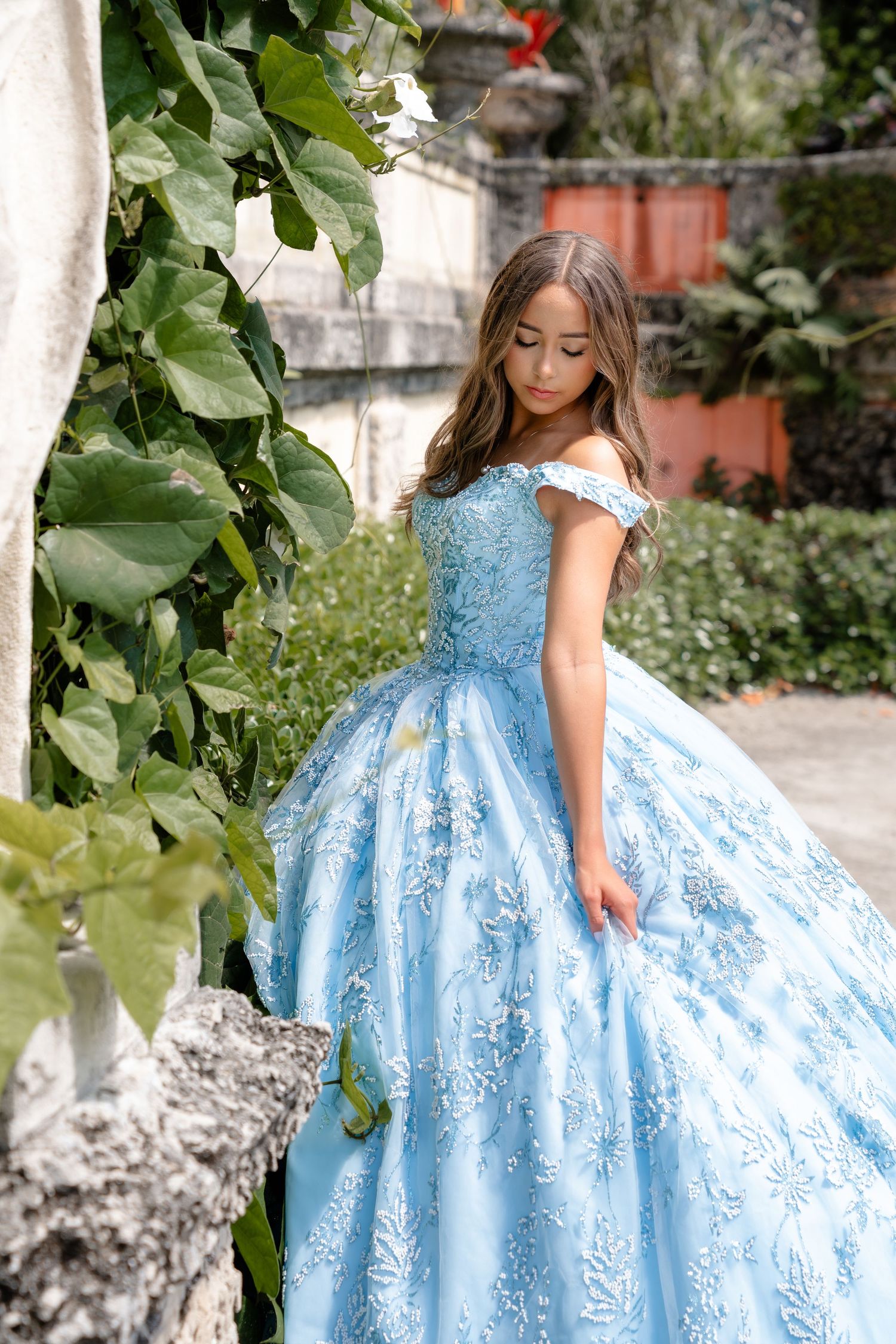 Young girl in light blue floral ball gown posing in lush garden setting.