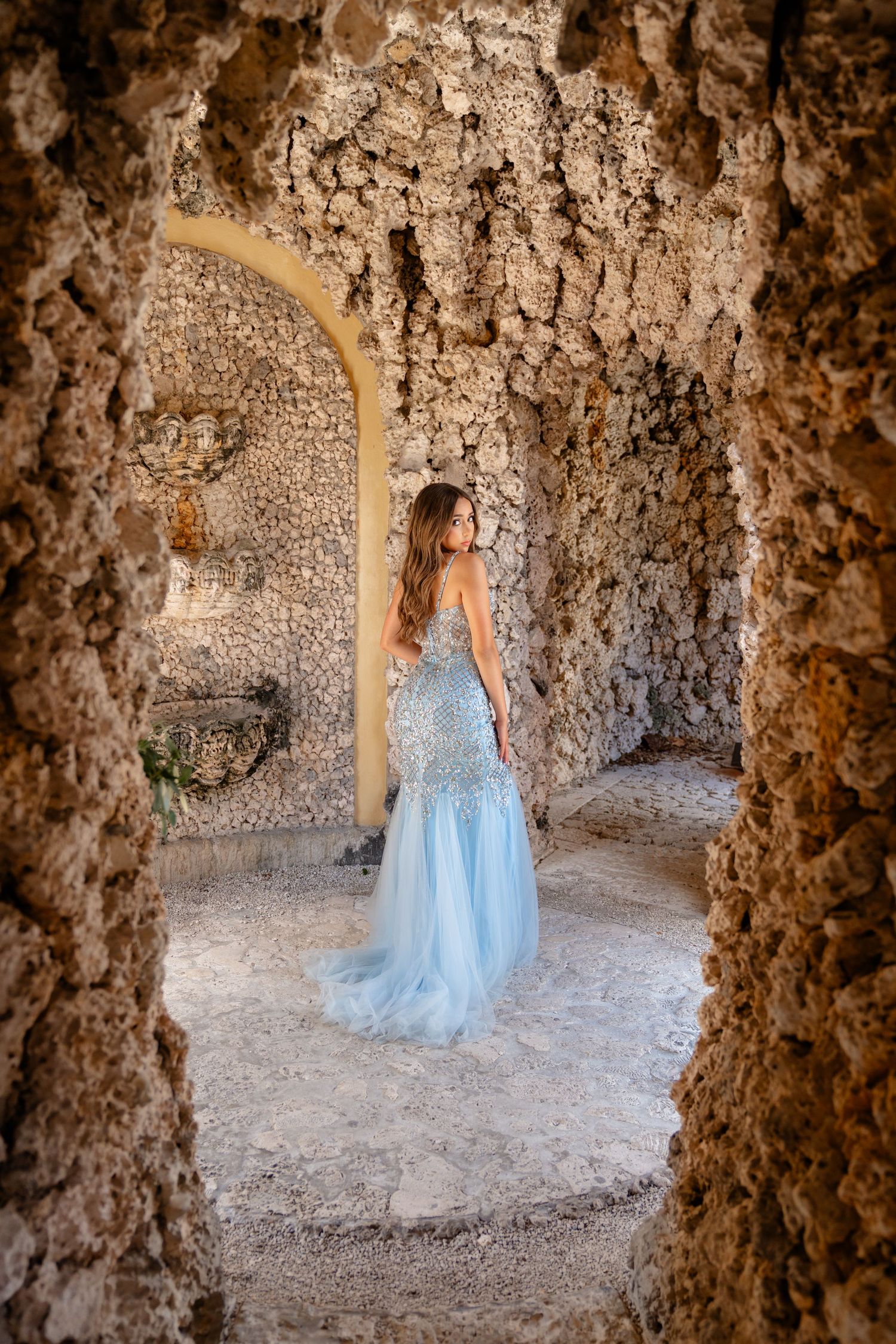 Woman in elegant blue gown standing in rustic stone grotto archway, long flowing dress with intricate beading details.