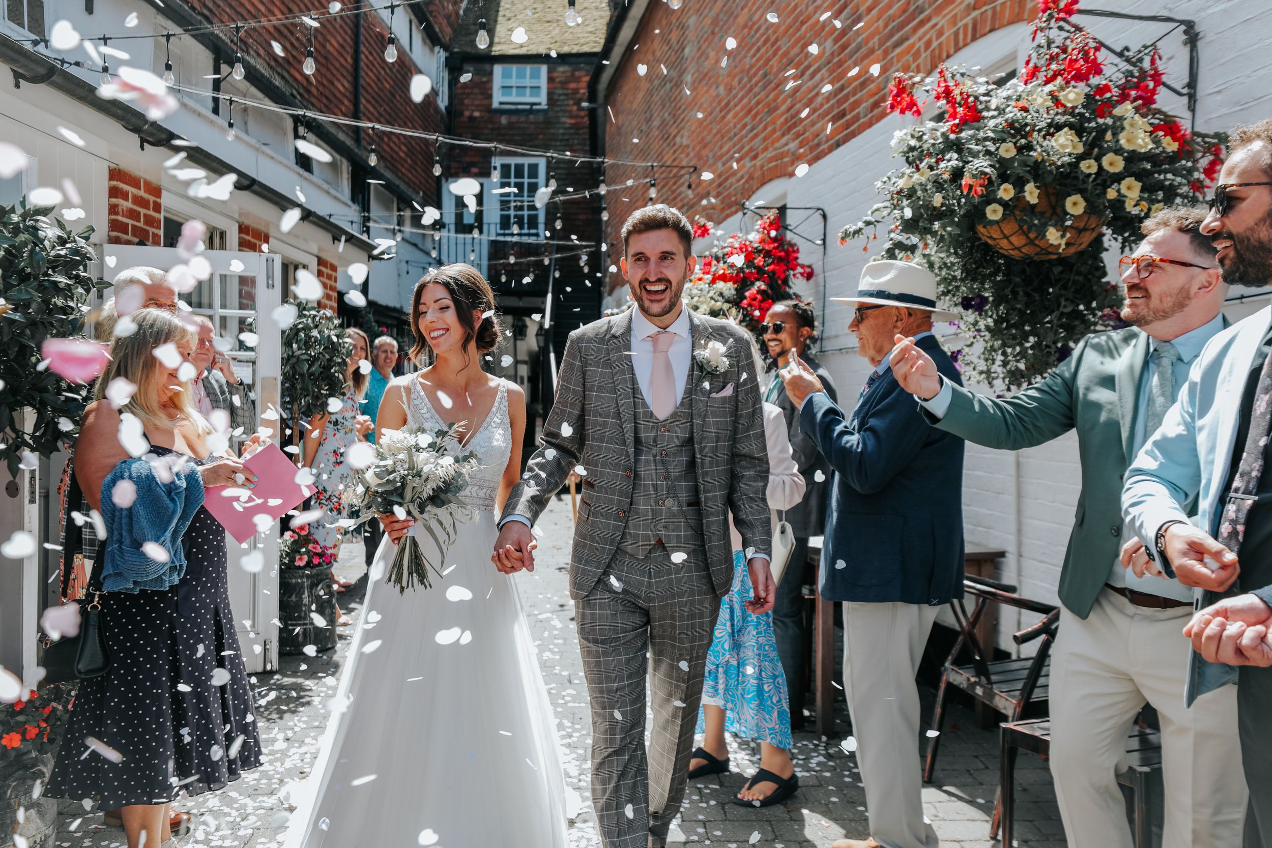 Newlyweds walk through shower of confetti with wedding guests on sunny street.