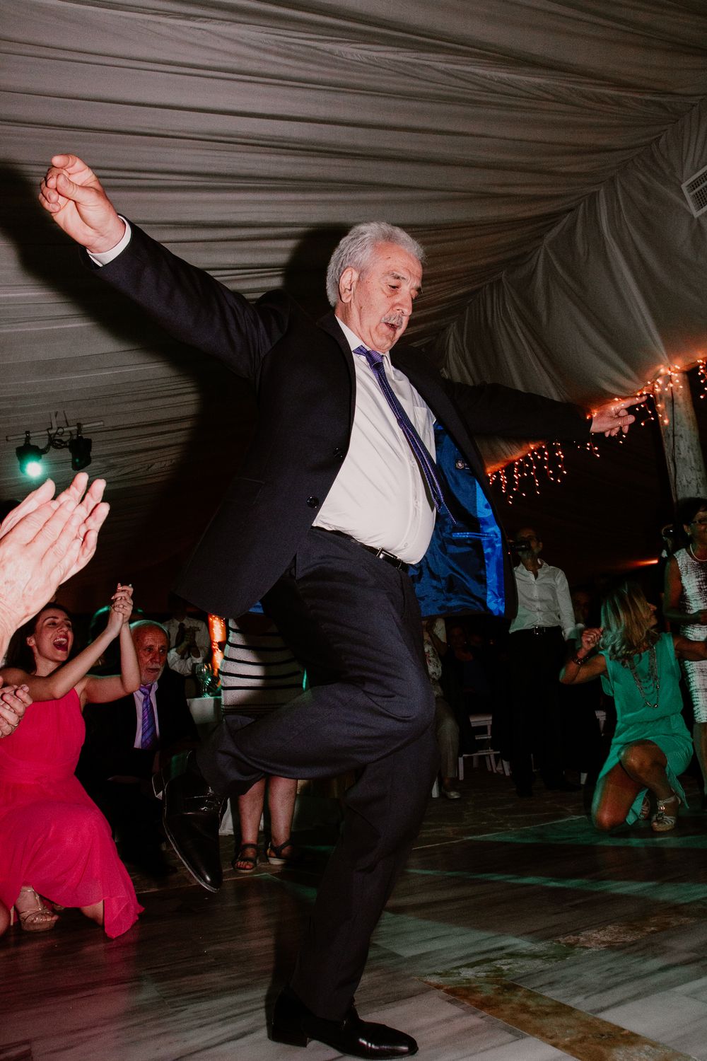 Energetic older man in suit with blue tie high-kicks on wedding reception dance floor as guests cheer.