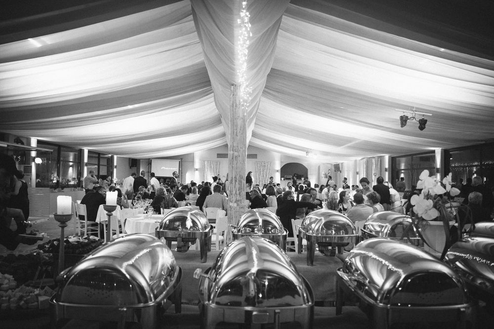 Black and white photo of an elegant wedding reception under draped ceiling with buffet chafing dishes in foreground.