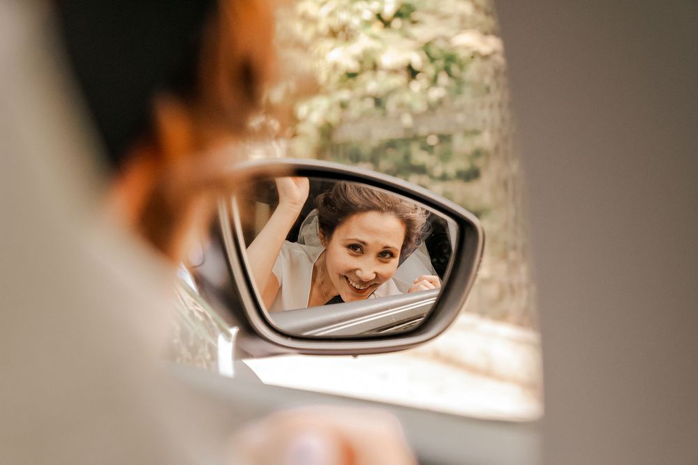 Smiling bride reflected in a car side mirror on a sunny day.